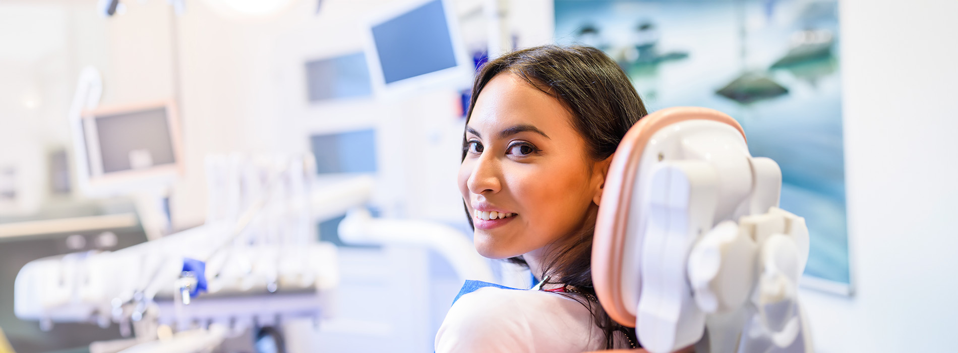 A person is seated in a dental chair, receiving care from a dental professional who stands behind them.